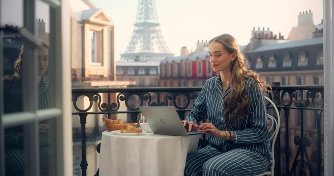 Woman Sits on a Parisian Balcony, Laptop Computer Open in Front of Her, Writing Content for Her Travel Blog. View of the Eiffel Tower and Morning Coffee Provides Inspiration for Her Creative Work