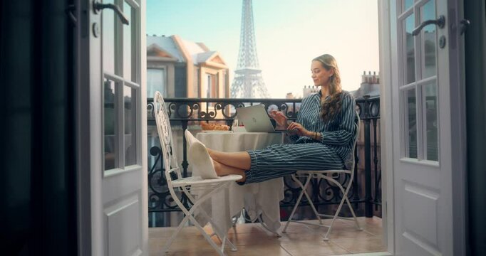 Woman in Pajamas Enjoying a Quiet Breakfast with Coffee while Using Her Laptop on a Balcony. She Casually Put Her Feet on a Chair and Shops Online as the Eiffel Tower Stands Tall in the Distance