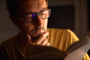 Bookworm reading literature book at night in dark room, his face is lit by desk lamp light