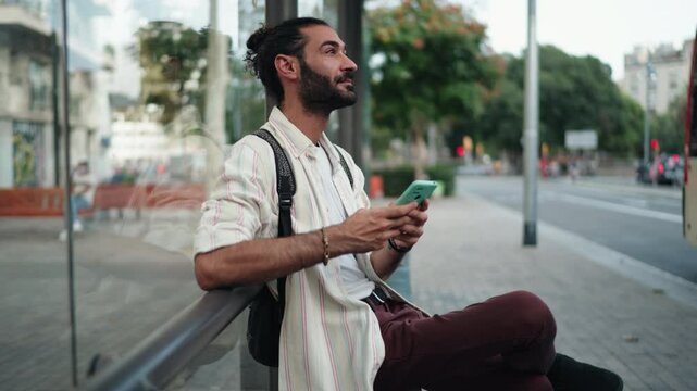 Hispanic man waiting at bus stop, checking public transport schedule on smartphone. Male tourist relaxed and prepared, using mobile app to stay informed about arrival times in urban setting
