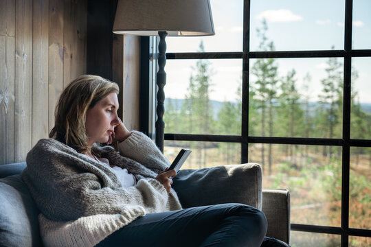 A woman sits comfortably on a couch in a cabin, engrossed in her e-reader book. Sunlight streams through large window, showcasing a beautiful forest landscape