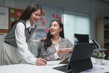 Two young businesswomen discuss the details of their planning, operations, and income during coffee breaks at their desks happily.