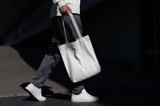 Man walking with a blank tote bag mockup template in an urban outdoor environment with sharp sunlit shadows. Isolated surface to place your design. 