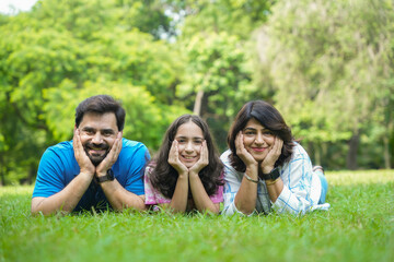 Happy young Indian family of three lying at summer park, embracing and smiling at camera, Parents with daughter enjoying quality time together. Togetherness.