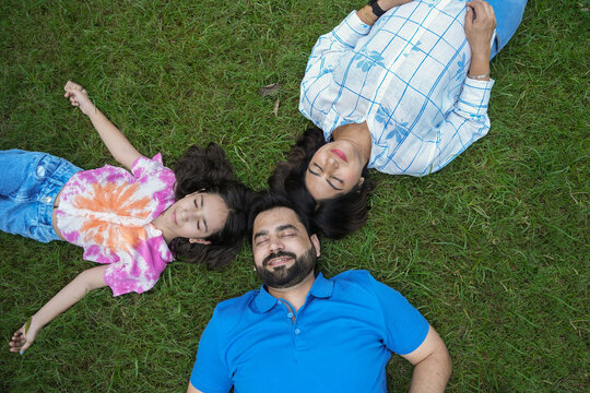 Top view of happy young indian family of three lying on grass in circle eyes closed. Parents with child daughter Relaxing, Togetherness. paren