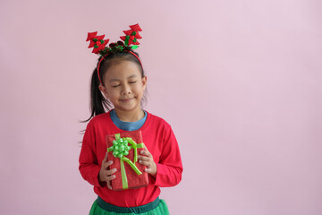 Portrait on a pink background of a cute little girl in a headband holding a happy red gift box with a beautiful green bow in various verses. Christmas Eve New Year and Winter holidays.