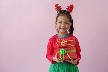 Portrait on a pink background of a cute little girl in a headband holding a happy red gift box with a beautiful green bow in various verses. Christmas Eve New Year and Winter holidays.