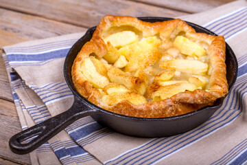 Homemade apple tart in a cast iron skillet, with slices of golden fruit and crisp puff pastry. The hot pan is resting on a tea towel over a rustic wood table.