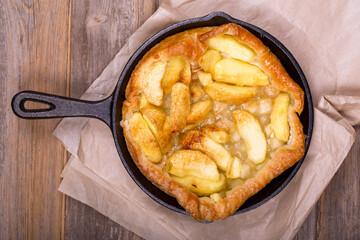 Homemade apple tart in a cast iron skillet, with slices of golden fruit and crisp puff pastry. The hot pan is resting on baking parchment over a rustic wood table. Overhead view.