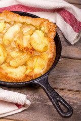 Homemade apple tart in a cast iron skillet, with slices of golden fruit and crisp puff pastry. The hot pan is resting on a tea towel over a rustic wood table.