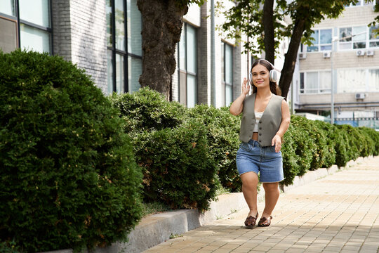 A cheerful brunette young woman with short stature strolls along a path lined with vibrant greenery, embracing the sunny day outdoors.