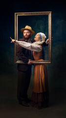 Couple, man and woman, dressed in retro clothing, posing with golden frame. Blending historical and modern themes. News, sales, announcement. Concept of art, eras comparison, creative vision, ad