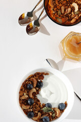Breakfast. Natural Greek Yoghurt with Freshly Baked Granola Served in White Bowl, Glass Jar of Honey, Spoons Arranged on White Table.