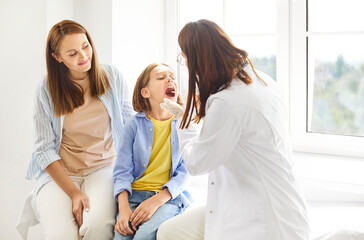 Fototapeta premium Female family pediatrician examining little girl patient's throat in medical clinic during checkup. Doctor otolaryngologist exam the oral cavity of a sick child sitting with her mother in the office.