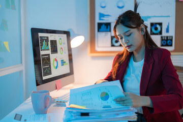 Businesswoman reviewing financial documents, working overtime in a dimly lit office with the glow...