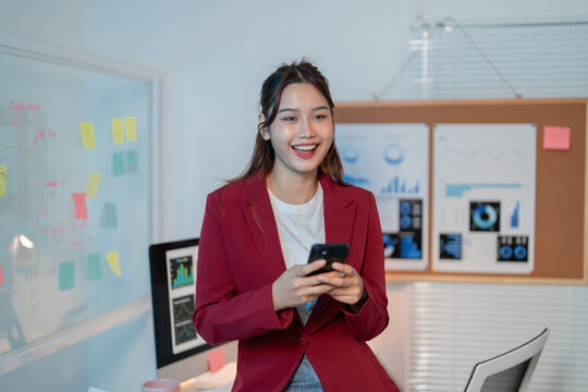 Confident young manager in modern office, smiling and using smartphone for business communication. Walls display charts symbolizing career success