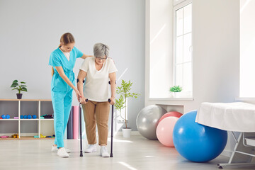 Young female caregiver, nurse helping elderly woman to walk with crutches during rehabilitation in...