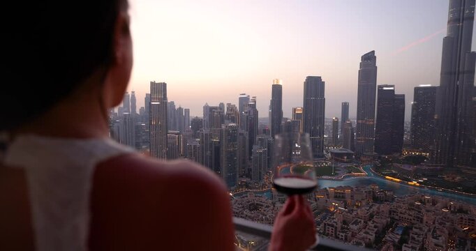 A beautiful tourist woman in a white dress holding a drink enjoys the evening view behind the modern skyline of Downtown Dubai, UAE