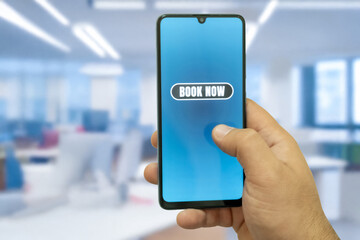 Close-up of the “Book Now” message on the smartphone screen. Online booking concept. Office background. Early booking message. 