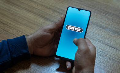 Close-up of the “Book Now” message on the smartphone screen. Online booking concept. Office background. Early booking message. 