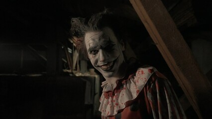 Crazy creepy killer man dressed as clown with makeup at attic in ruined house looks at camera,...