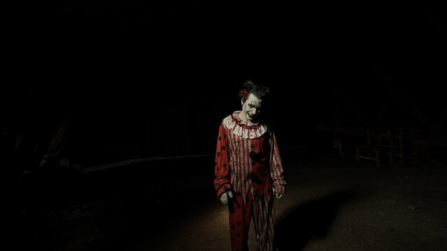 Crazy killer man dressed as clown with creepy makeup standing looking at camera at attic in ruined house, Halloween thriller concept.