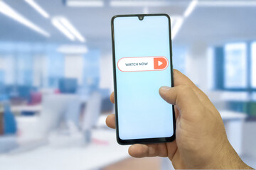 “Watch Now” message on close-up smartphone screen. Watching online video. Office background. Video streaming platform, video advertising and streaming concept.