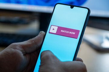 “Watch Now” message on close-up smartphone screen. Watching online video. Office background. Video streaming platform, video advertising and streaming concept.