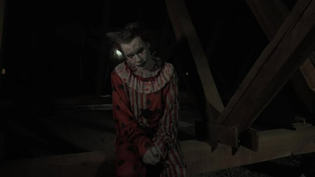 Crazy creepy killer man dressed as clown with makeup at attic in ruined house sits on wooden beam swinging legs sad face. Halloween thriller concept.