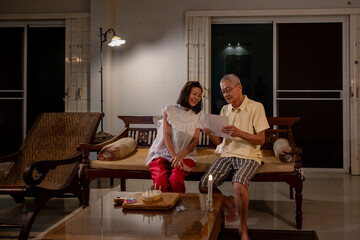 Senior asian couple is happily celebrating birthday together at home while exchanging best wish card for love, relationship, bonding, longevity and marriage