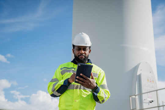 A man in a yellow and black safety vest is looking at a tablet. He is wearing a hard hat and safety glasses. electrical, engineer, environment, energy, clean, sustainable, wind turbine, nature