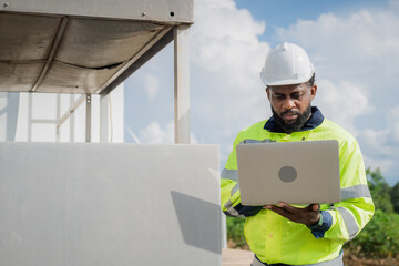 A man wearing a yellow and black safety vest is looking at a laptop. He is wearing a hard hat and safety glasses. electrical, engineer, environment, energy, clean, sustainable, wind turbine, nature