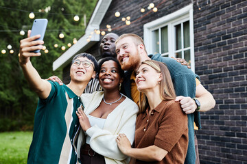 Waist up portrait of multiethnic group of people taking selfie photo together outdoors by house decorated with fairy lights