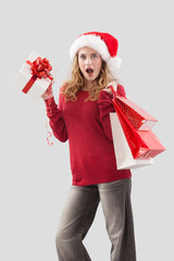 amazed merry Christmas woman wearing Santa hat holds gift box and red shopping bags. Image for sale offer promotions, online shopping, advertising banner, poster, or gift card, isolated background