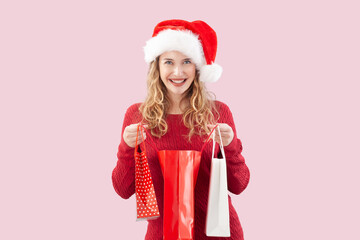 Smiling blonde young woman opening a Christmas present shopping bag while wearing a Santa hat, isolated on a pink background. For sale and offer promotions, online shopping adv banner, or gift card