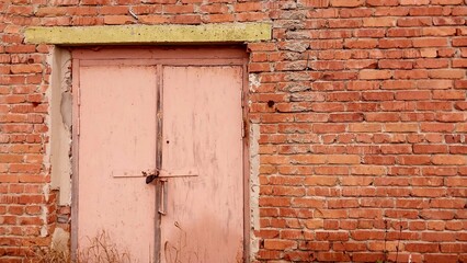 a section of a brick wall with a closed wooden door. The door is painted in a faded pink color and appears to be old and weathered. It is secured with a metal latch and has a horizontal wooden beam ac