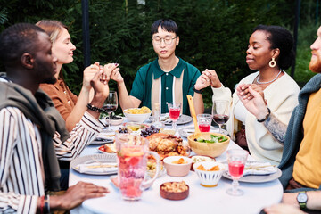 Multicultural group of people celebrating Thanksgiving together and holding hands sitting at dinner table outdoors shot with flash