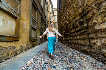 Holidays in Italy. Blonde woman wearing beige sweater, turquoise skirt walking with hands spread out along cobblestone narrow street in old town of Bergamo in autumn. Back view. Sightseeing old city.