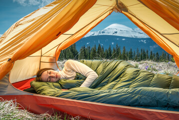 Young woman sleeping under a down blanket in a yellow tent in the morning mountains.