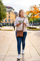 City break. Portrait of beautiful middle aged woman wearing jeans, beige sweater, sunglasses walking with city map on square and looking around during sightseeing european city on sunny autumn day.