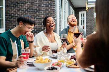 Candid scene of group of adult friends enjoying drinks at dinner table outdoors and laughing...