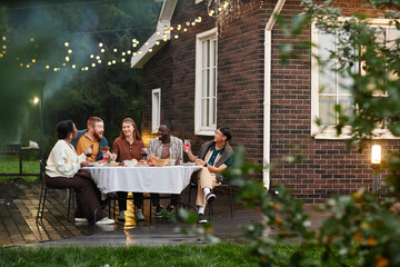 Wide angle shot of diverse group of adult people enjoying dinner party together at outdoor patio by brick house copy space