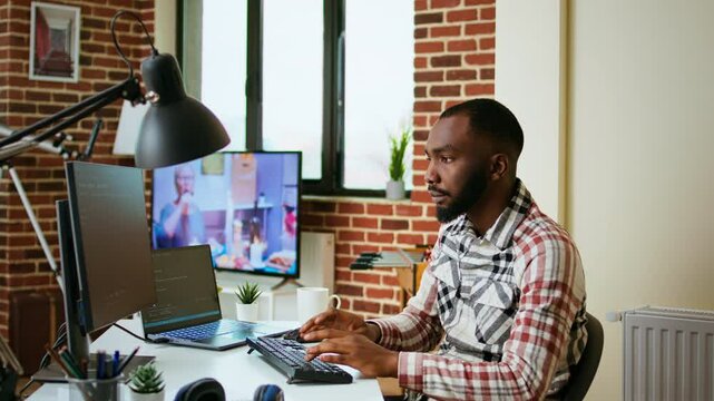 Male coder works from home typing code on a laptop in a modern living room. African american IT engineer using programming language and cloud computing for software development. Camera A.