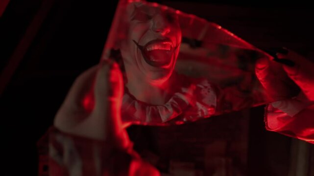 Crazy creepy killer man dressed as clown with makeup at attic in ruined house holding mirror shard looks in it crazy smiling. Halloween thriller concept.