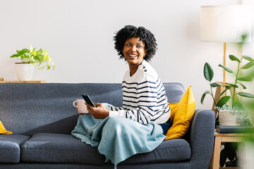 Young Latin American woman enjoying a cozy winter day at home, wrapped in a blanket on the couch...