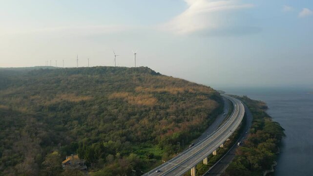 Aerial view of a highway winding through a hilly, forested landscape with wind turbines in the distance, near a coastline.
