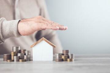 Businessman making a protective hand gesture over a stack of coins and a house model. Image...