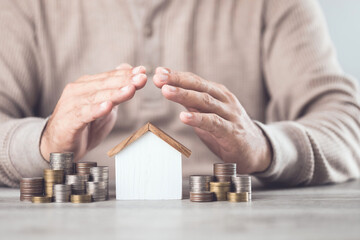 Businessman making a protective hand gesture over a stack of coins and a house model. Image represents the concept of asset protection, emphasizing safeguarding financial investments and property