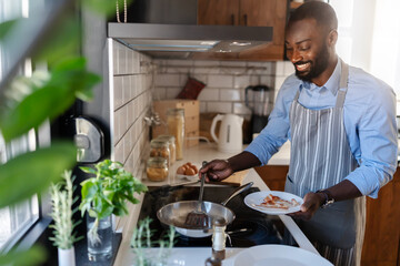 A young, smiling African-American man is frying bacon for breakfast in his modern kitchen. He enjoys a peaceful morning while preparing his meal in a bright and stylish space.