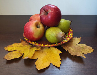 Red apples and ripe pears lie on a plate, near them there are maple leaves